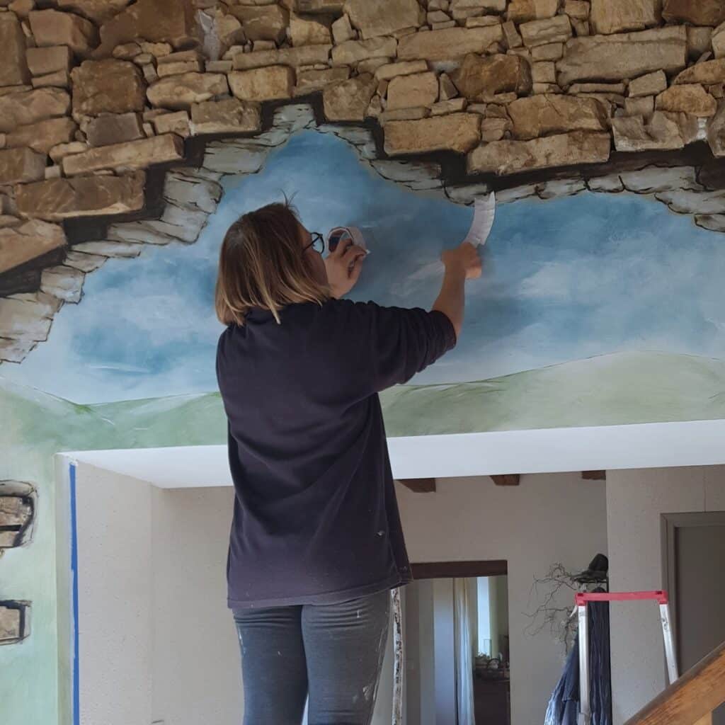 Création d'une fresque murale trompe-l'œil ciel et pierre Artiste créant un trompe-l'œil de ciel nuageux sous une arche en pierres sèches.