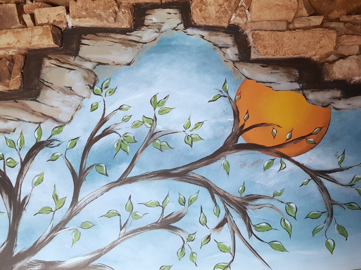 Mural Arbre et Soleil à travers un Mur de Briques Fresque murale d'un arbre aux feuilles vertes devant un soleil orange, vue à travers une brèche dans un mur de briques.