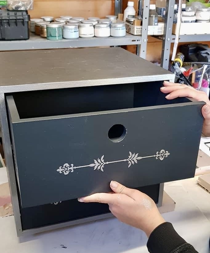 Rénovation de meuble avec peinture et pochoir argenté Mains tenant un tiroir gris foncé avec motif au pochoir argenté sur un petit meuble en cours de rénovation DIY.
