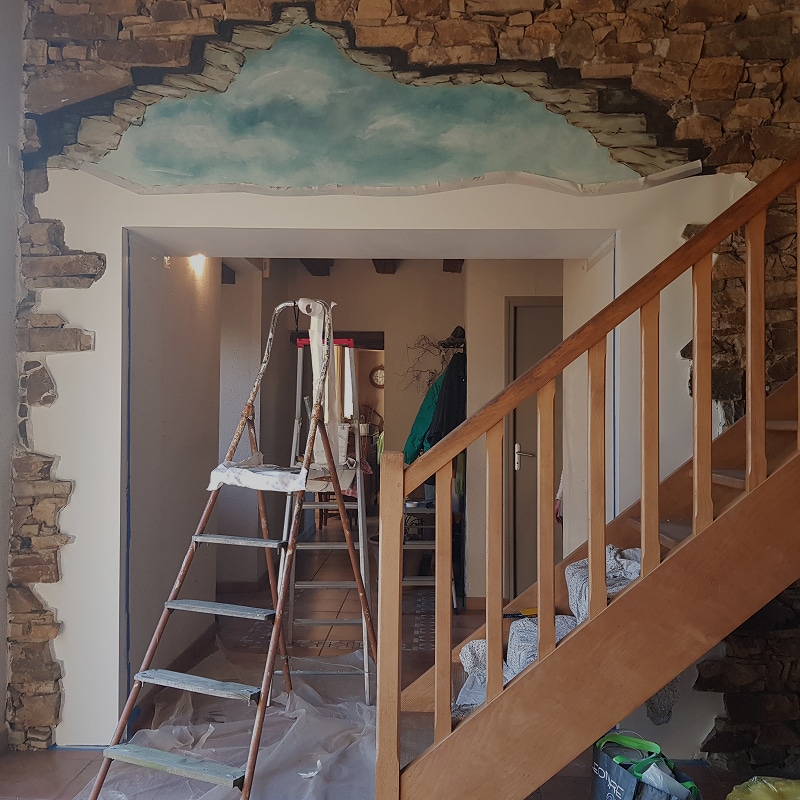 Rénovation Intérieure : Fresque Ciel et Mur de Pierres Fresque de ciel bleu en trompe-l'œil sur un mur de pierres en rénovation avec une échelle et un escalier en bois.
