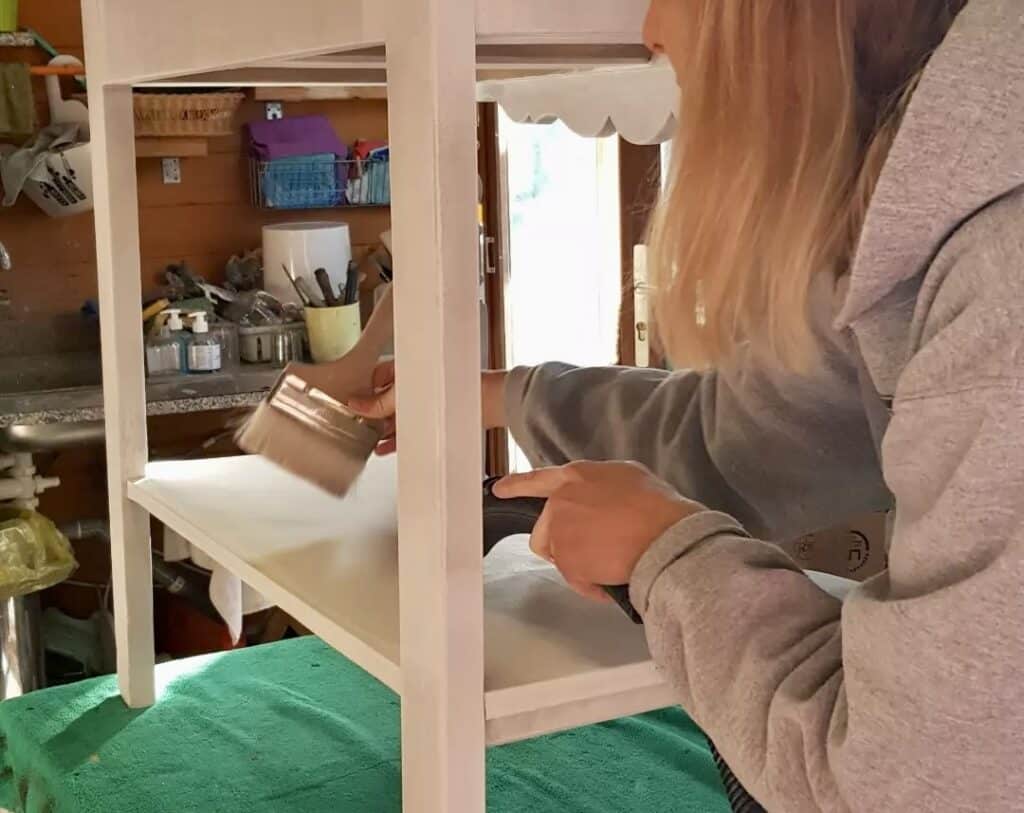 Restaurer et peindre un meuble en bois blanc - DIY Déco Femme peignant une étagère en bois blanc avec un pinceau et un pistolet thermique dans son atelier DIY.