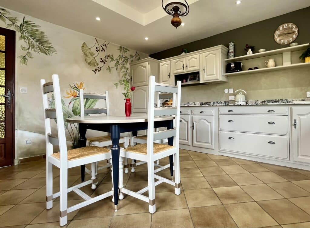Cuisine rustique avec chaises blanches, table noire, murale florale et horloge sur mur vert olive.
