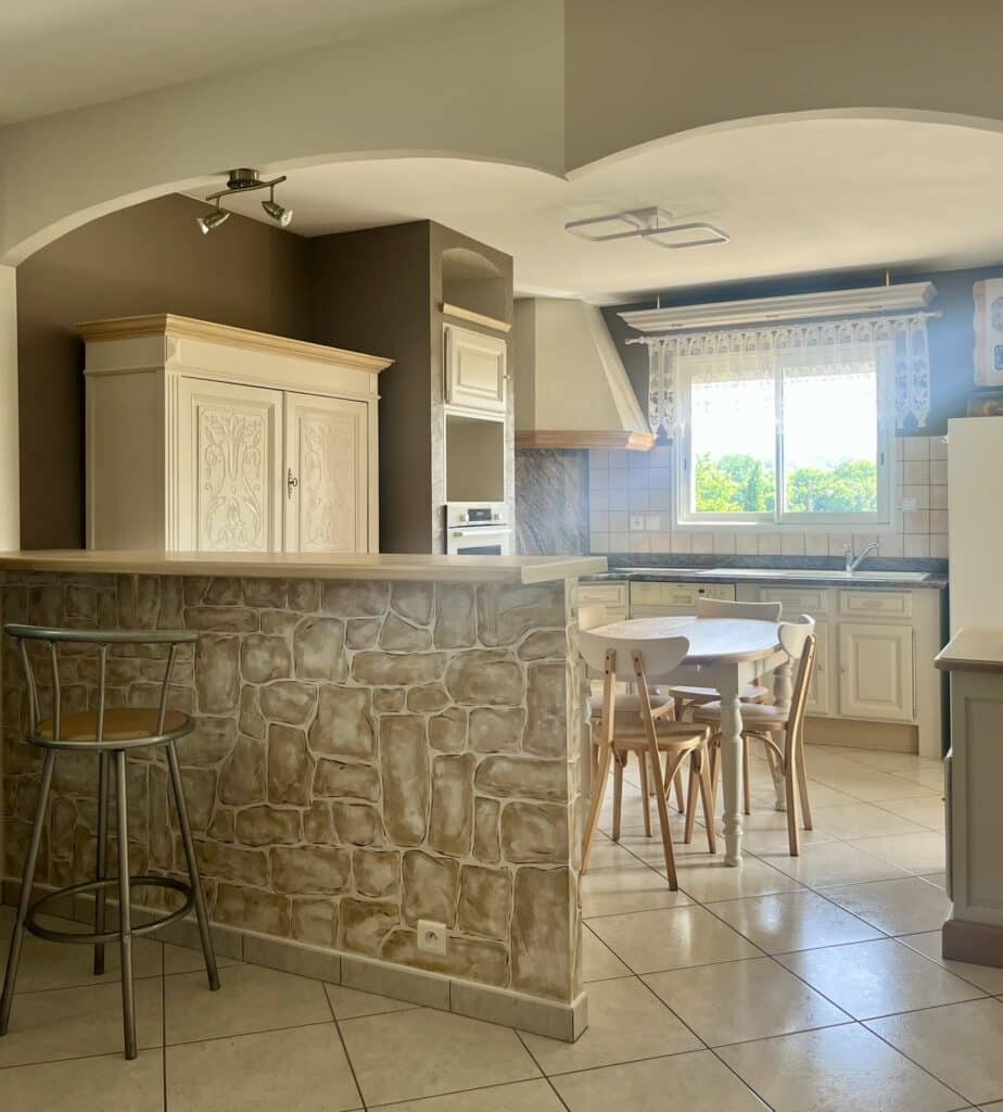 Cuisine rustique lumineuse avec bar en pierre, table bois et armoire sculptée décorative.