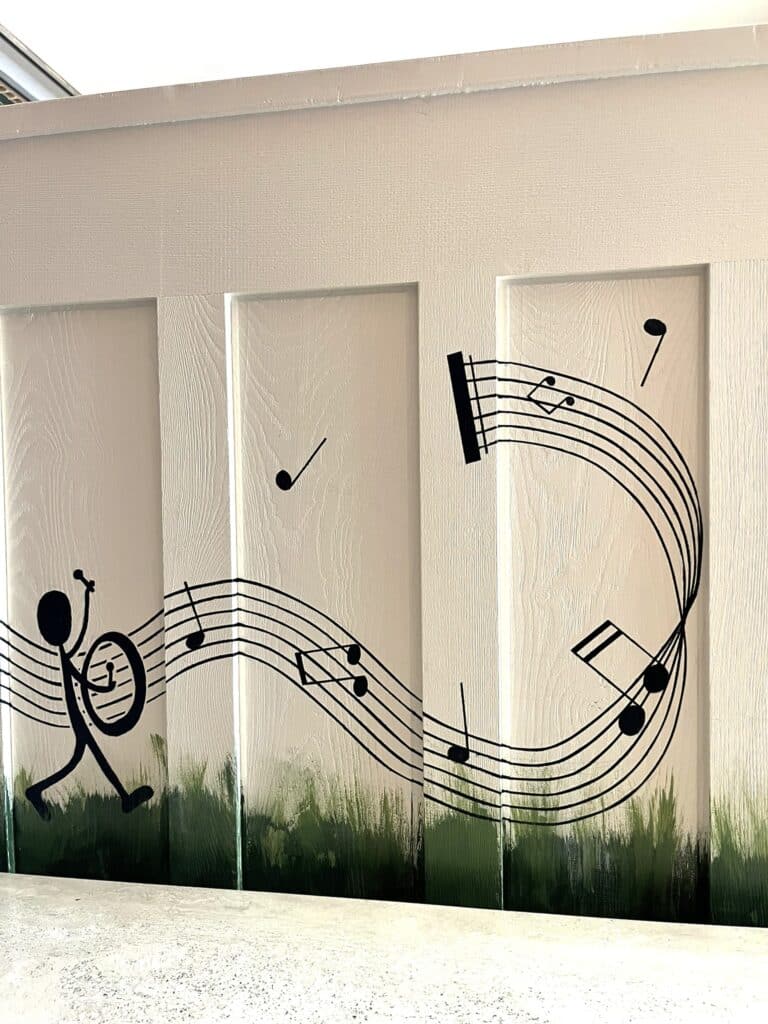 Mural d'un musicien bâton, tambour et portée de notes noires sur fond blanc avec herbe peinte.