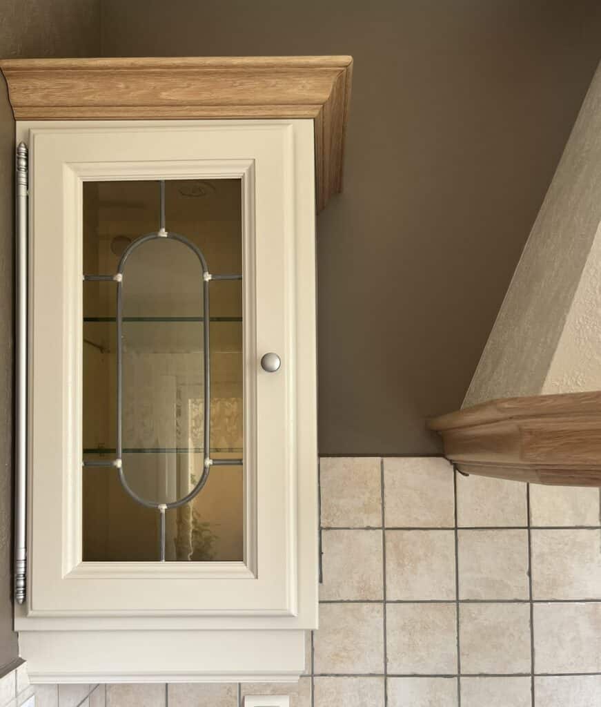 Placard de cuisine crème avec porte vitrée en plomb, corniche en chêne massif et carrelage beige.