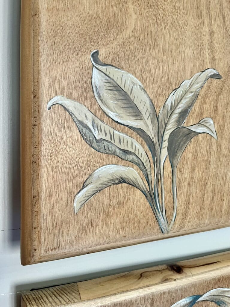 Peinture de feuilles blanches et grises sur un panneau de bois naturel au grain visible, décor mural botanique.